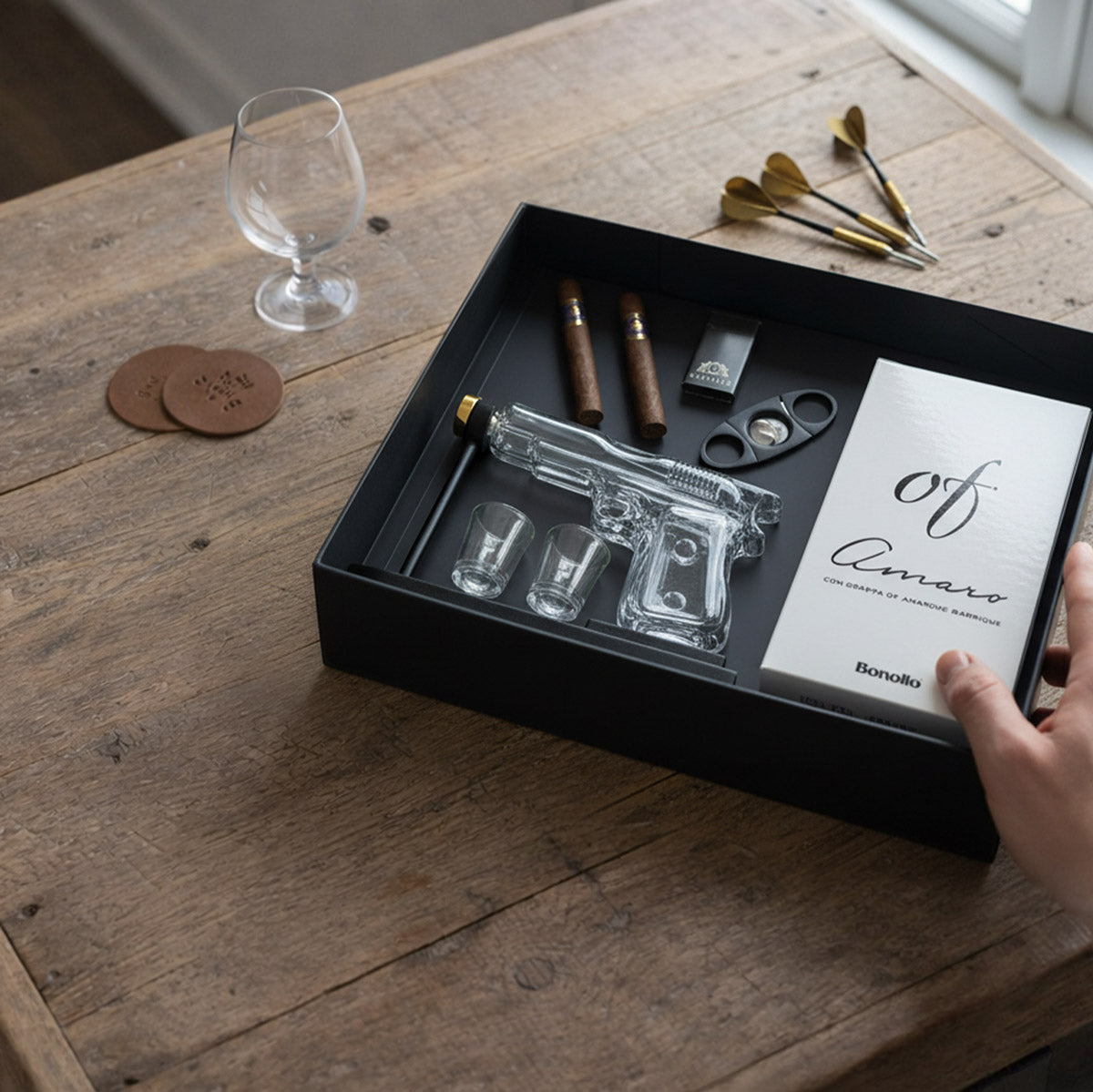 A gift box including a clear gun-shaped decanter, shot glasses, cigars, cigar lighter and cutter, and a bottle of alcohol, on a wooden table with a glass, coasters, and darts in the background